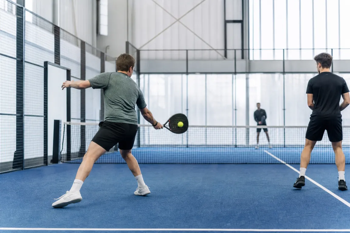 Padelspelare mitt i ett grundslag nära nätet på en bana med glasväggar