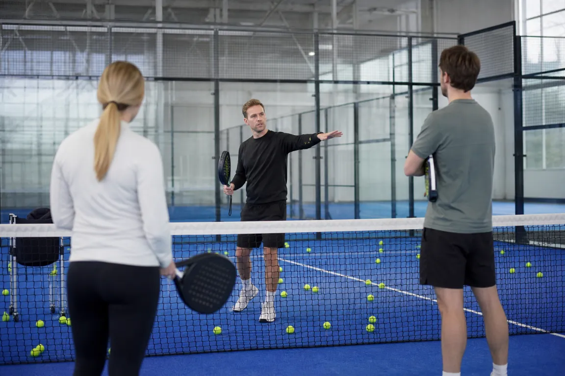 Tränare som instruerar två spelare vid nätet på en padelbana med bollar utspridda