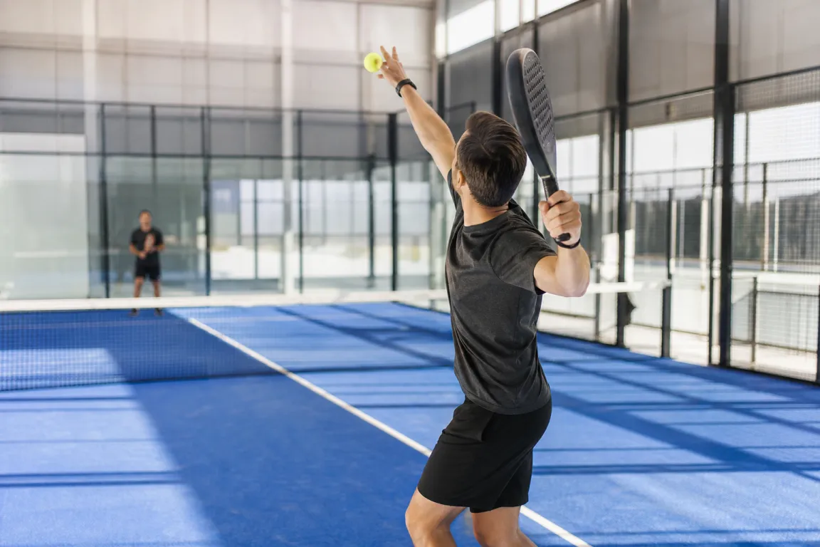 Padelspelare i serveposition vid baslinjen med glasväggar och galler synliga runt banan